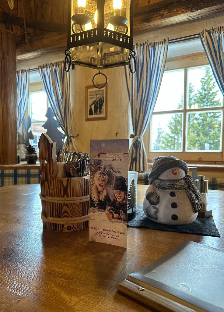 Ein gemütlicher hölzerner Esstisch mit Besteck, Schneemann-Dekoration und Speisekarte, der an einem Fenster mit gestreiften Vorhängen steht.
